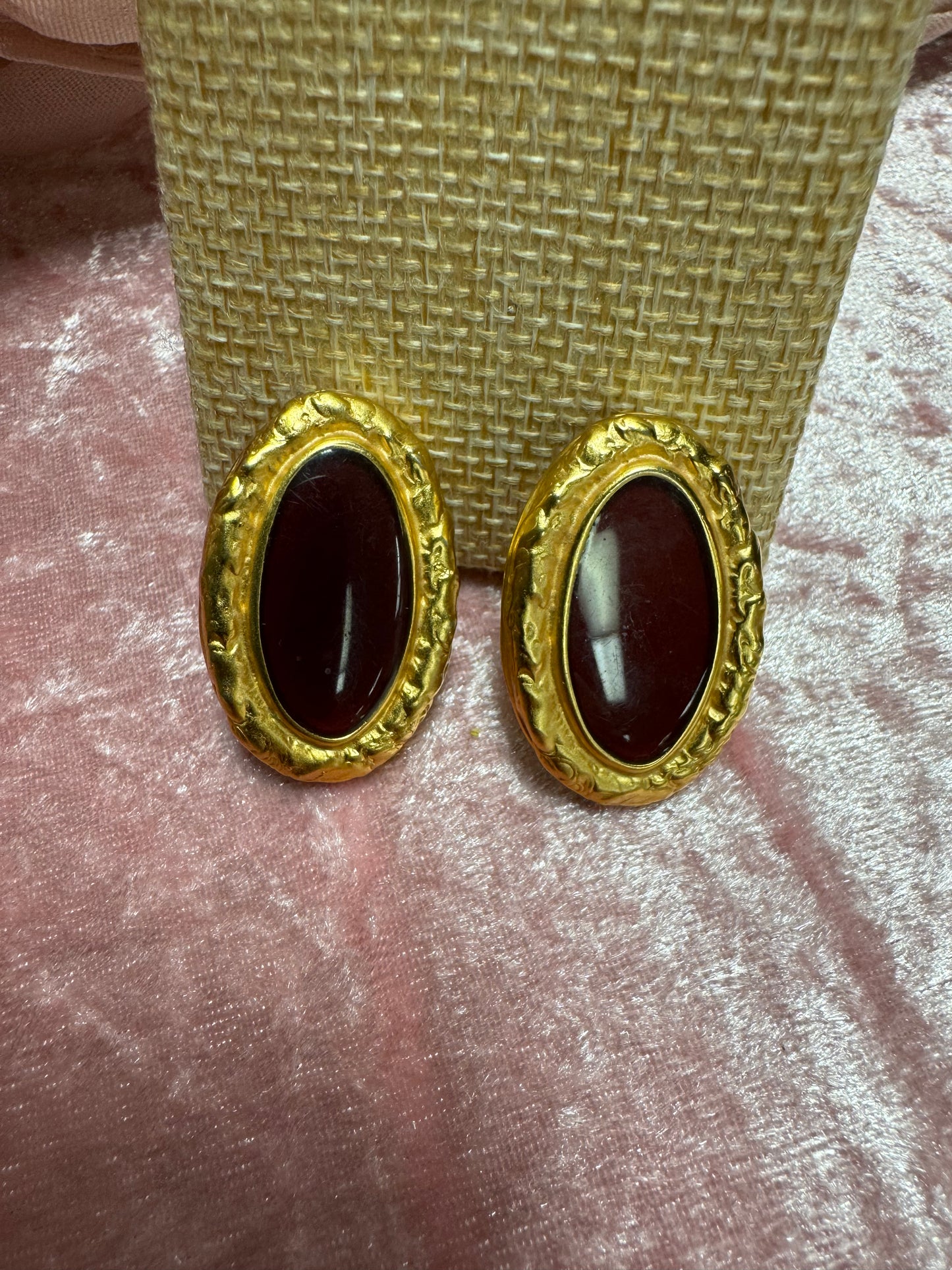 Vintage, gold-tone button earrings featuring a textured rope-style border surrounding a dark red, oval cabochon stone