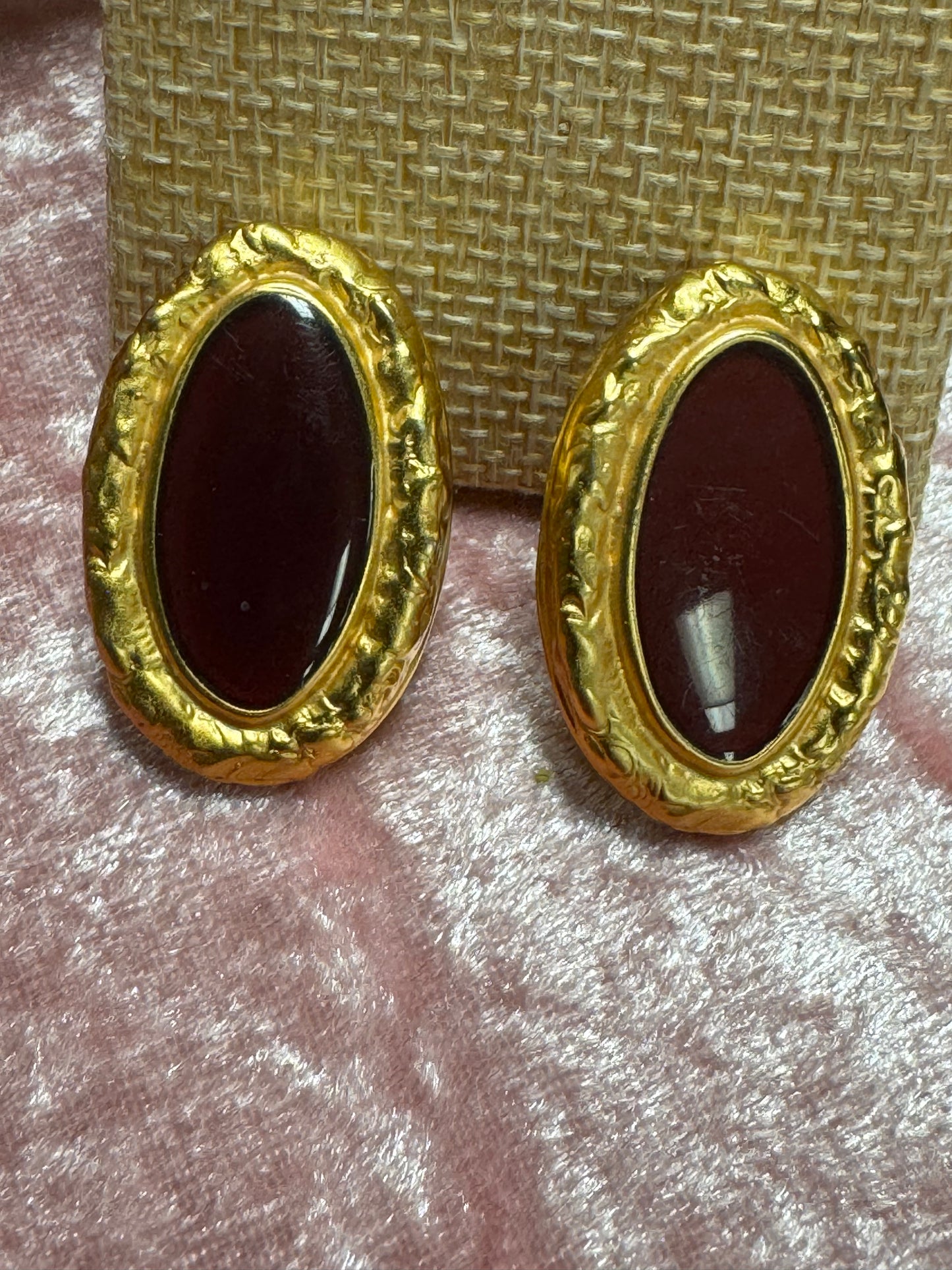 Vintage, gold-tone button earrings featuring a textured rope-style border surrounding a dark red, oval cabochon stone